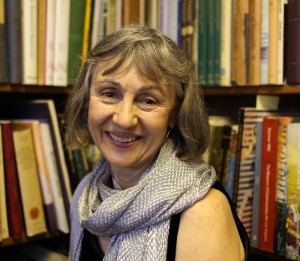 A photo of Alice Elliott, an older woman with light skin and shoulder-length grey hair wearing a black tank top and scarf, smiling widely at the camera, sitting in front of a bookshelf.