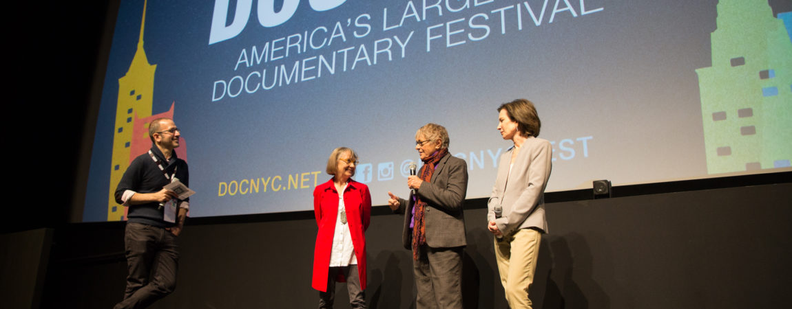 Alice, a white woman with a red blazer over a white shirt, with orange round glasses and a brown bob, is standing on stage in front of a large projector that says "Doc NYU America's Largest Documentary Festival". 3 other people are on stage with her.