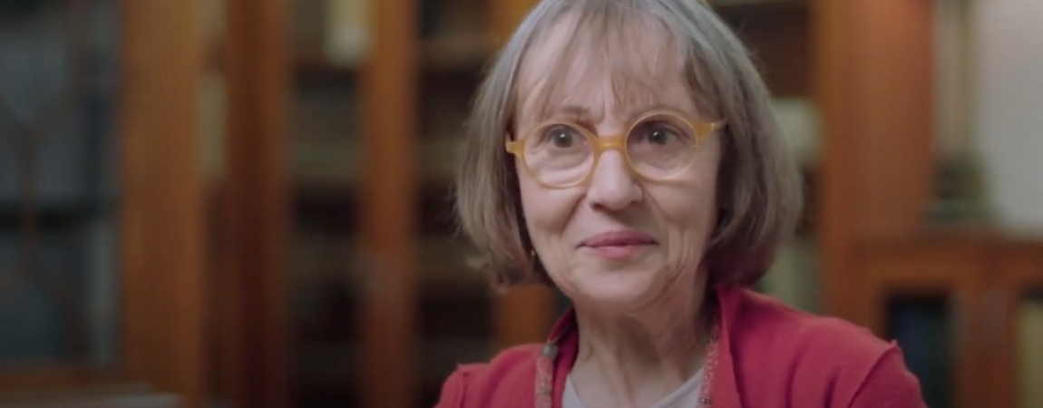 A photo of Alice, a white woman with a brown and grey bob, orange round glasses, and a red cardigan looking at someone behind the camera.