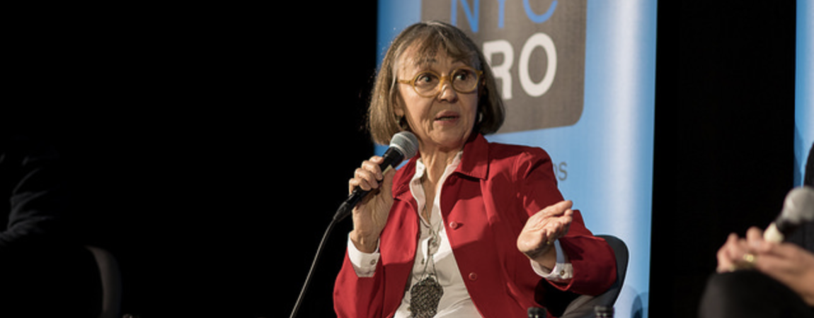 A photo of Alice, a white woman in a red blazer and white shirt with a brown bob, holding a microphone on stage while sitting in a chair. She is talking to someone off screen.